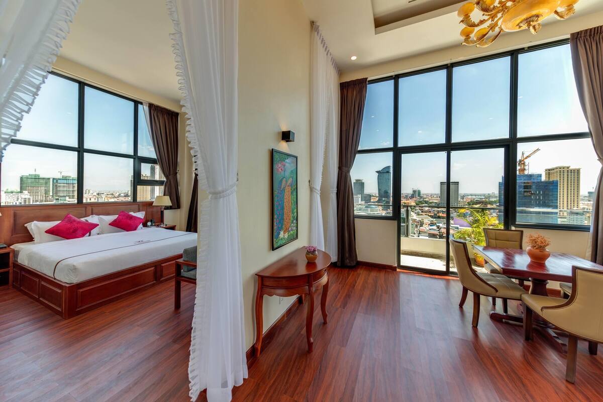 Una habitación de hotel con una cama grande, una mesa de comedor y vistas a la ciudad a través de amplios ventanales.