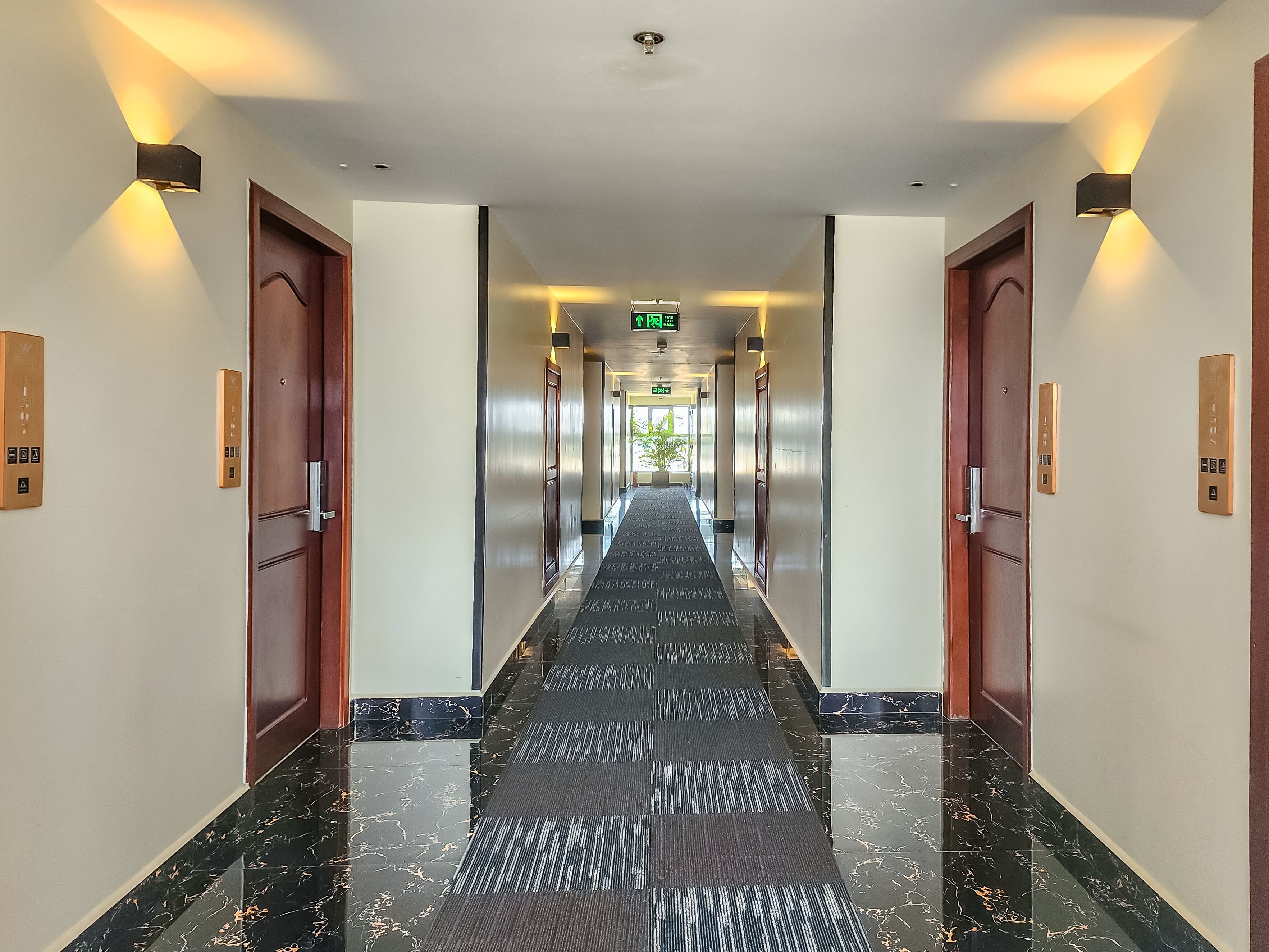 A hotel corridor with a black and white patterned floor, wooden doors, and wall-mounted light fixtures.