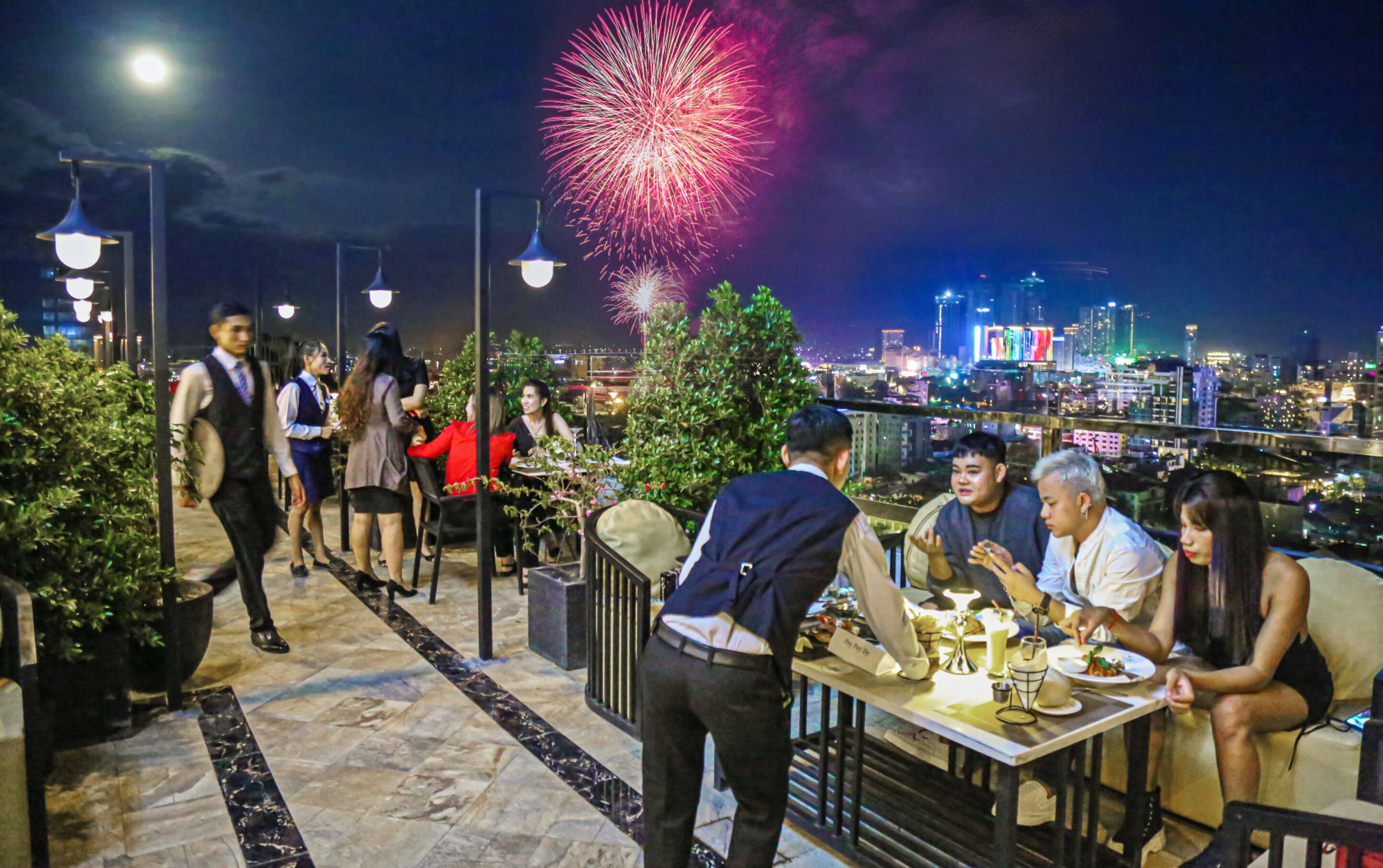 A rooftop restaurant with a city view, outdoor seating, and a waiter serving guests.