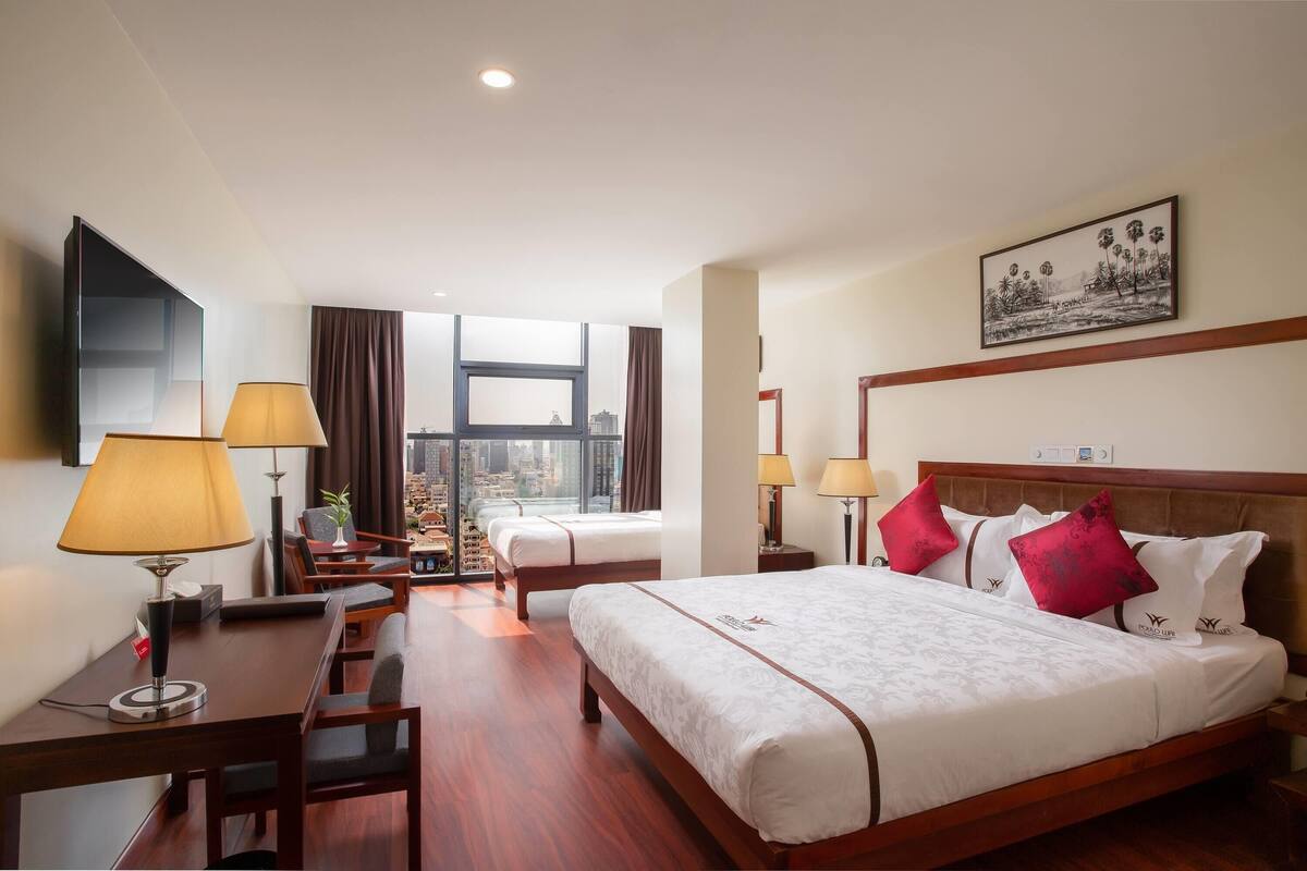 Habitación de hotel con una cama grande, dos mesitas de noche con lámparas, un escritorio con silla, un ventanal con vista a la ciudad y una obra de arte colgada en la pared.