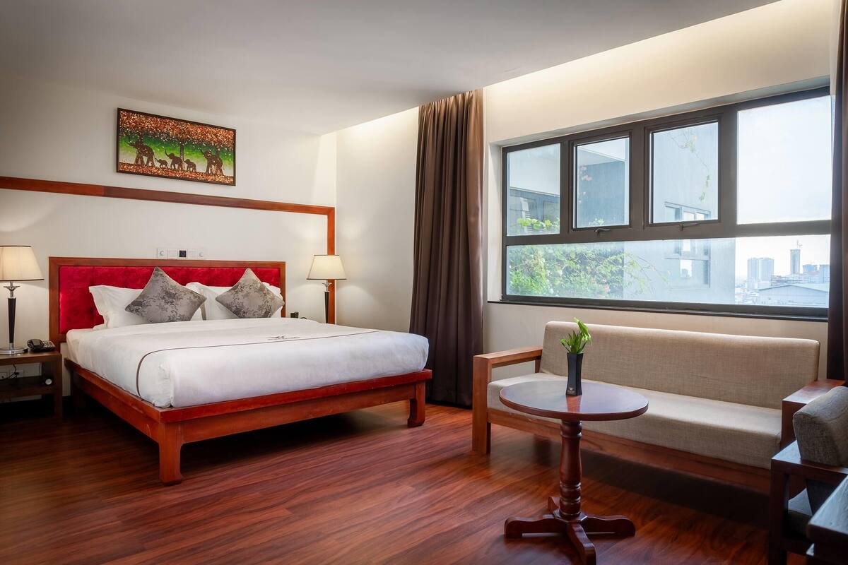 Una habitación de hotel con una cama de madera, un sofá, una mesita y un ventanal con vistas a edificios.