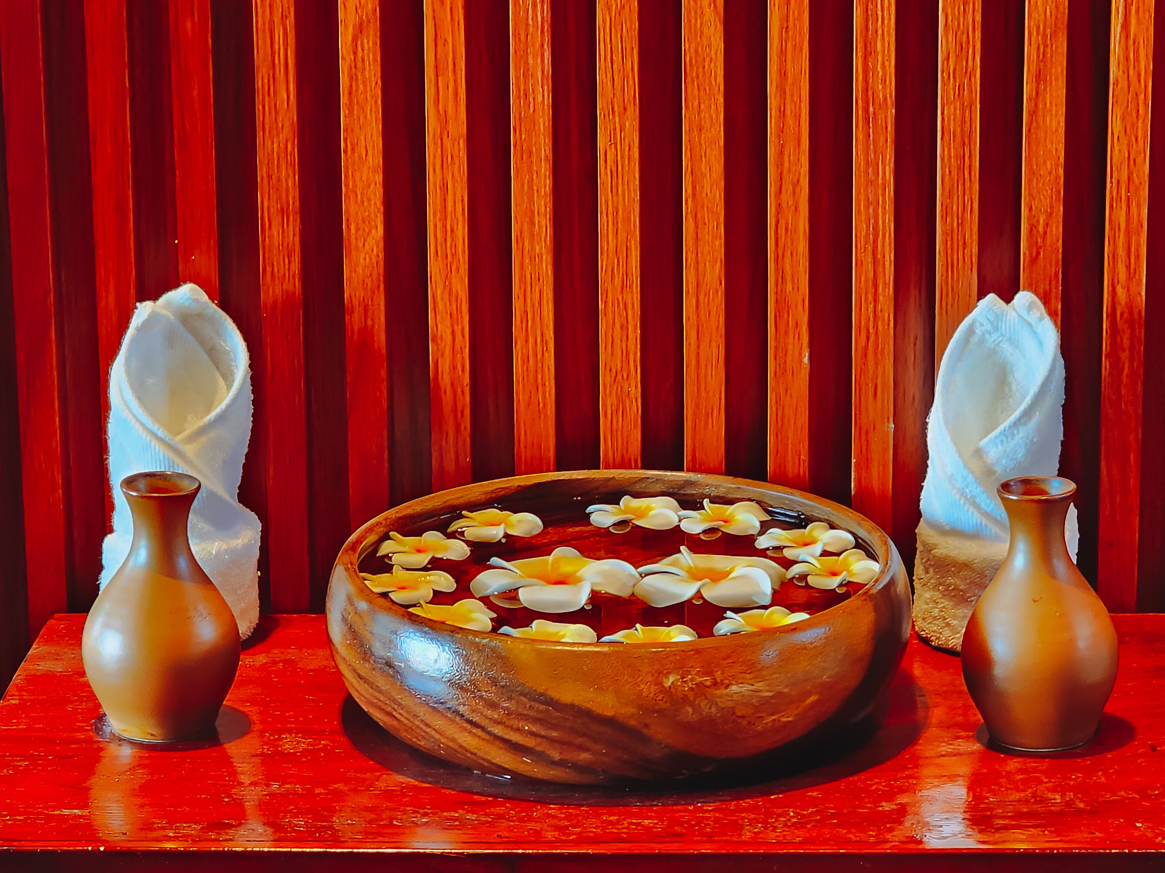 A wooden bowl filled with water and flower petals, placed on a red surface with two small vases on either side.