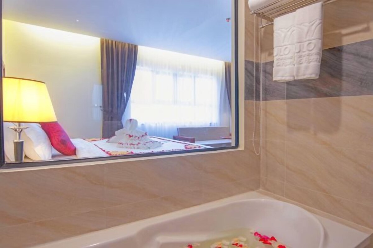 A bathroom with a bathtub, a mirror reflecting a room with a bed, a lamp, and a window with curtains.