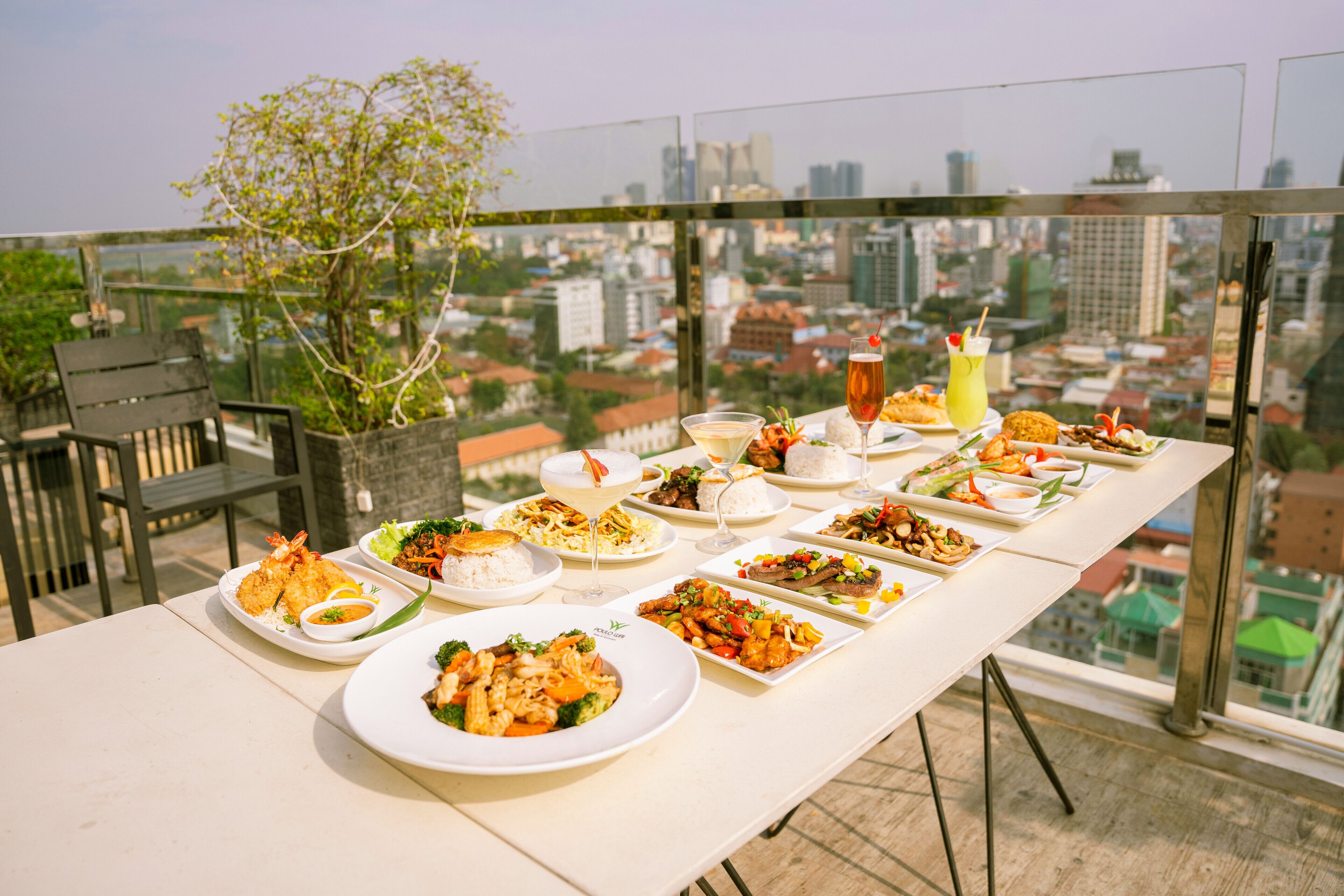 A table set with various dishes and drinks on a balcony with a city view.