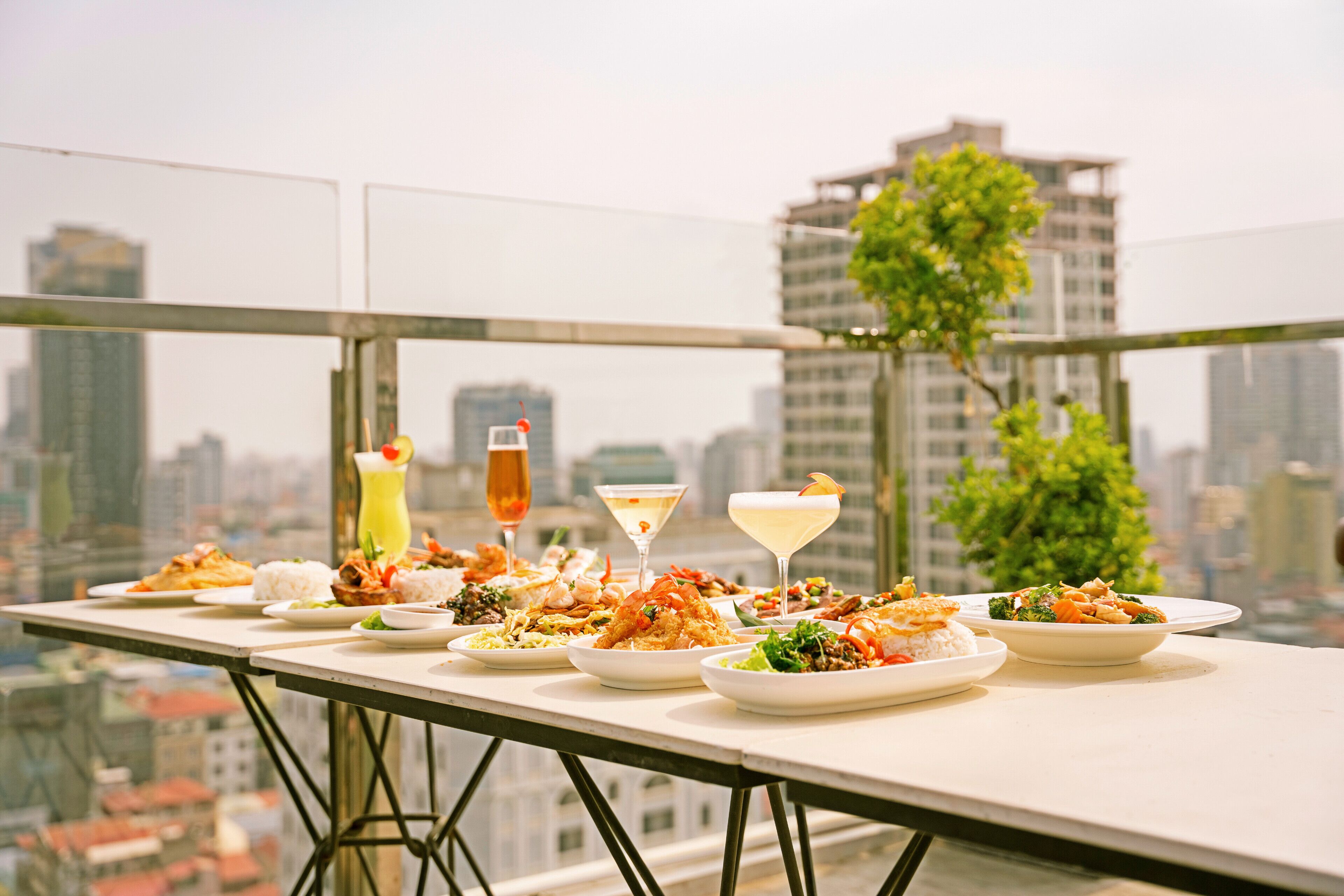 A dining table with various dishes and drinks on a balcony with a city view.
