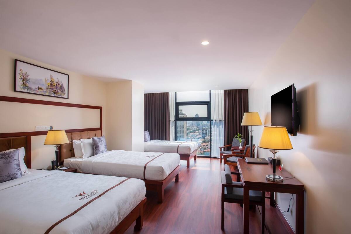 Habitación de hotel con dos camas, un escritorio con lámpara, televisión y ventana grande con vista a la ciudad.
