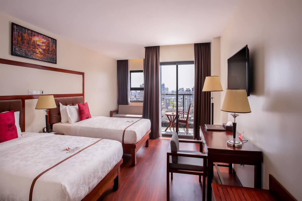 Habitación de hotel con dos camas, un escritorio, una silla, un televisor y un ventanal con vistas a la ciudad.