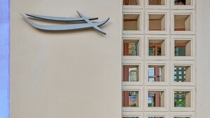 A building facade with a series of square windows, a decorative metal piece resembling a boat, and some greenery in the foreground. - Barabara Budget Satun (La-ngu)
