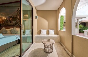 Una habitación con cama, un separador de vidrio, una mesa redonda de mimbre y un banco con cojines.