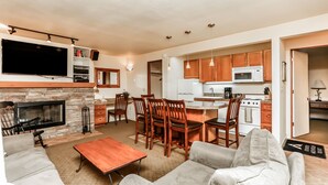 A modern living room with a fireplace, a dining area with wooden chairs, a kitchen with wooden cabinets, and a flat-screen TV mounted above the fireplace. - Stunning Views, Closest to Mt. Bachelor, Resort Amenities & Year-Round Fun! (Bend)