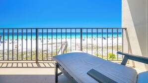Ein Balkon mit einer Matratze und Blick auf einen Strand mit Sonnenschirmen und Menschen.
