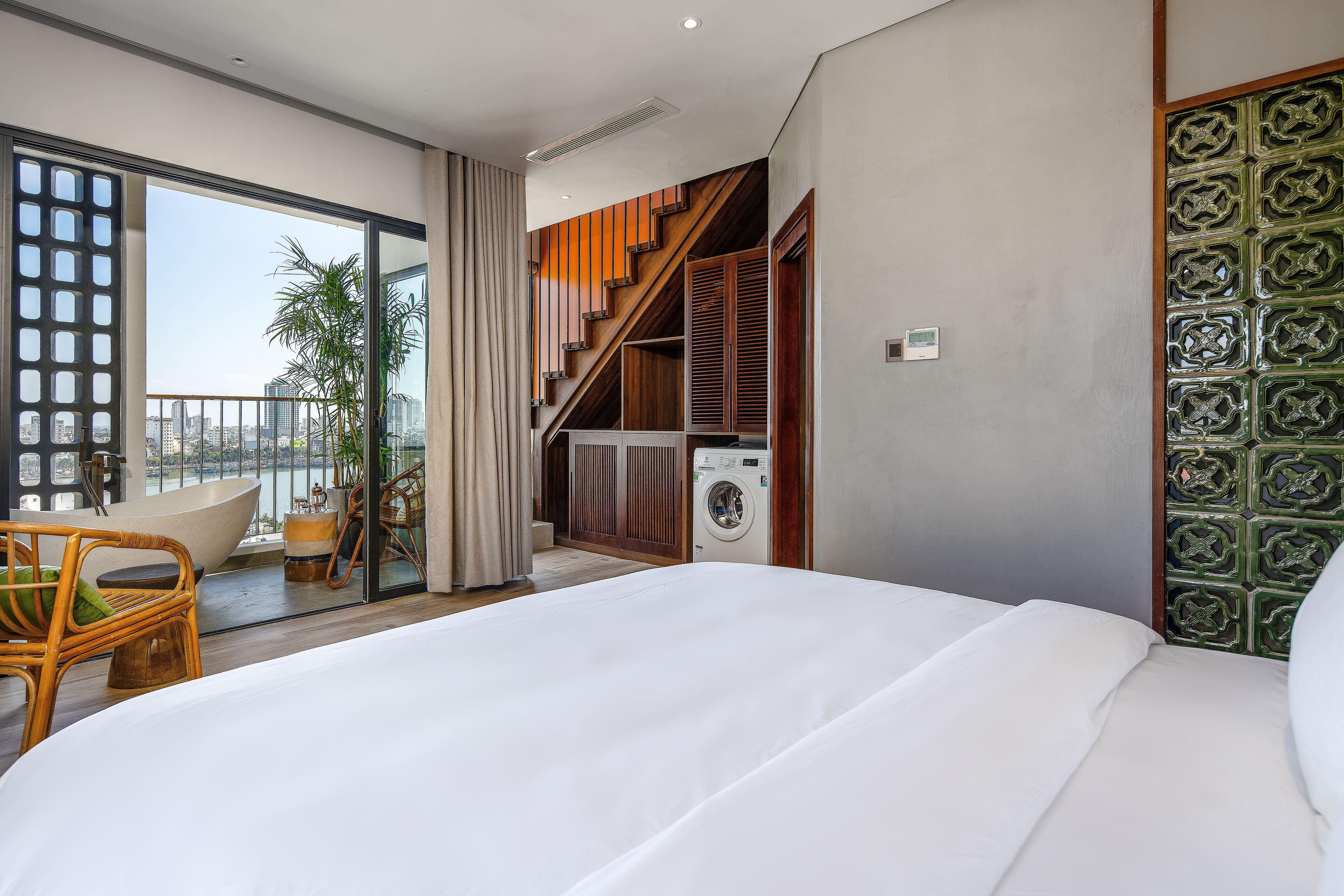 Una habitación de hotel moderna con una cama grande, un balcón con vistas a la ciudad, una lavadora y una zona de comedor con mobiliario de mimbre.