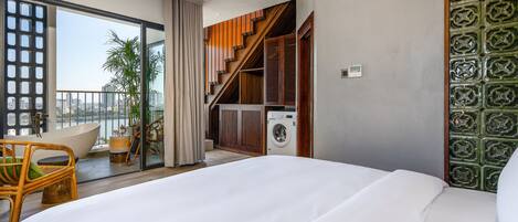 Una habitación de hotel moderna con una cama grande, un balcón con vistas a la ciudad, una lavadora y una zona de comedor con mobiliario de mimbre.