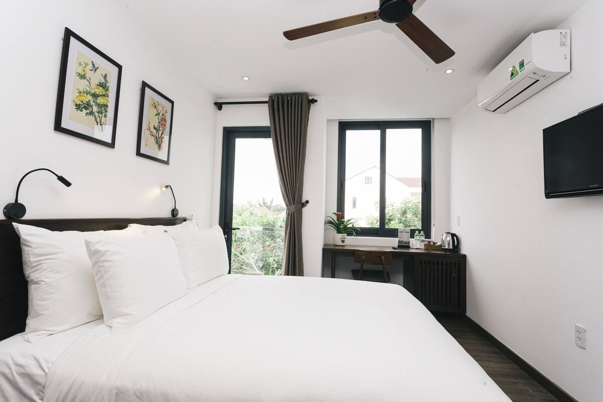 A hotel room with a bed, a desk, a chair, a ceiling fan, a window with curtains, and a mounted TV.