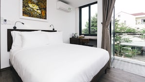 A bedroom with a large bed, a desk, a chair, a window with a view of the outside, and a painting on the wall. - Le Petit Villa Hoi An (Da Nang)