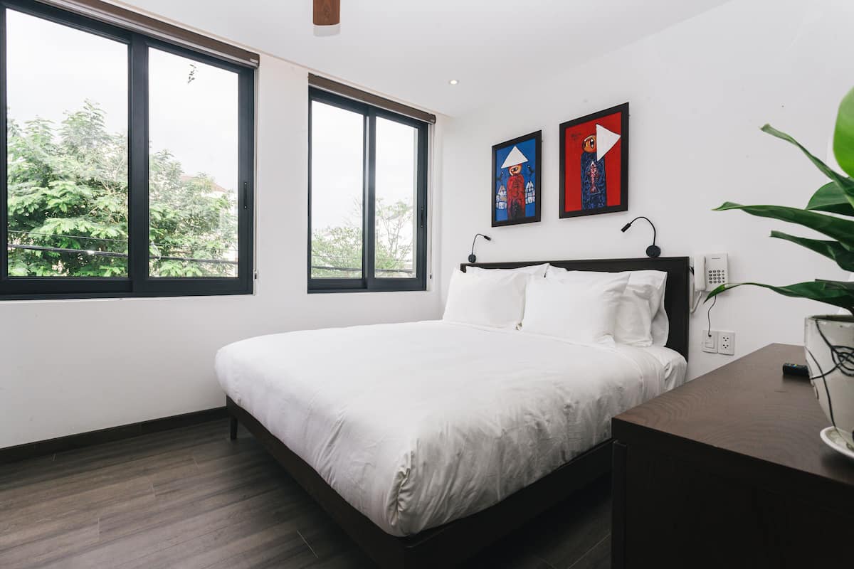 A bedroom with a large window, a bed, a bedside table, and two wall-mounted artworks.