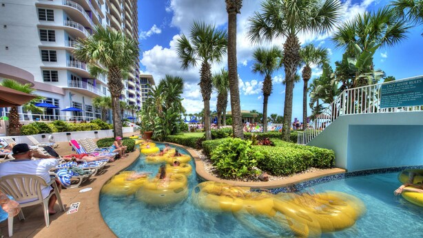 A pool area with multiple people in yellow inflatable tubes, surrounded by palm trees and a hotel building.