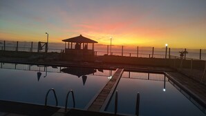 En swimmingpool med en pavillon og liggestole, omgivet af et hegn og udsigt til solnedgangen over vandet.