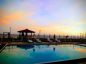 A swimming pool with lounge chairs and a gazebo overlooking the ocean at sunset.