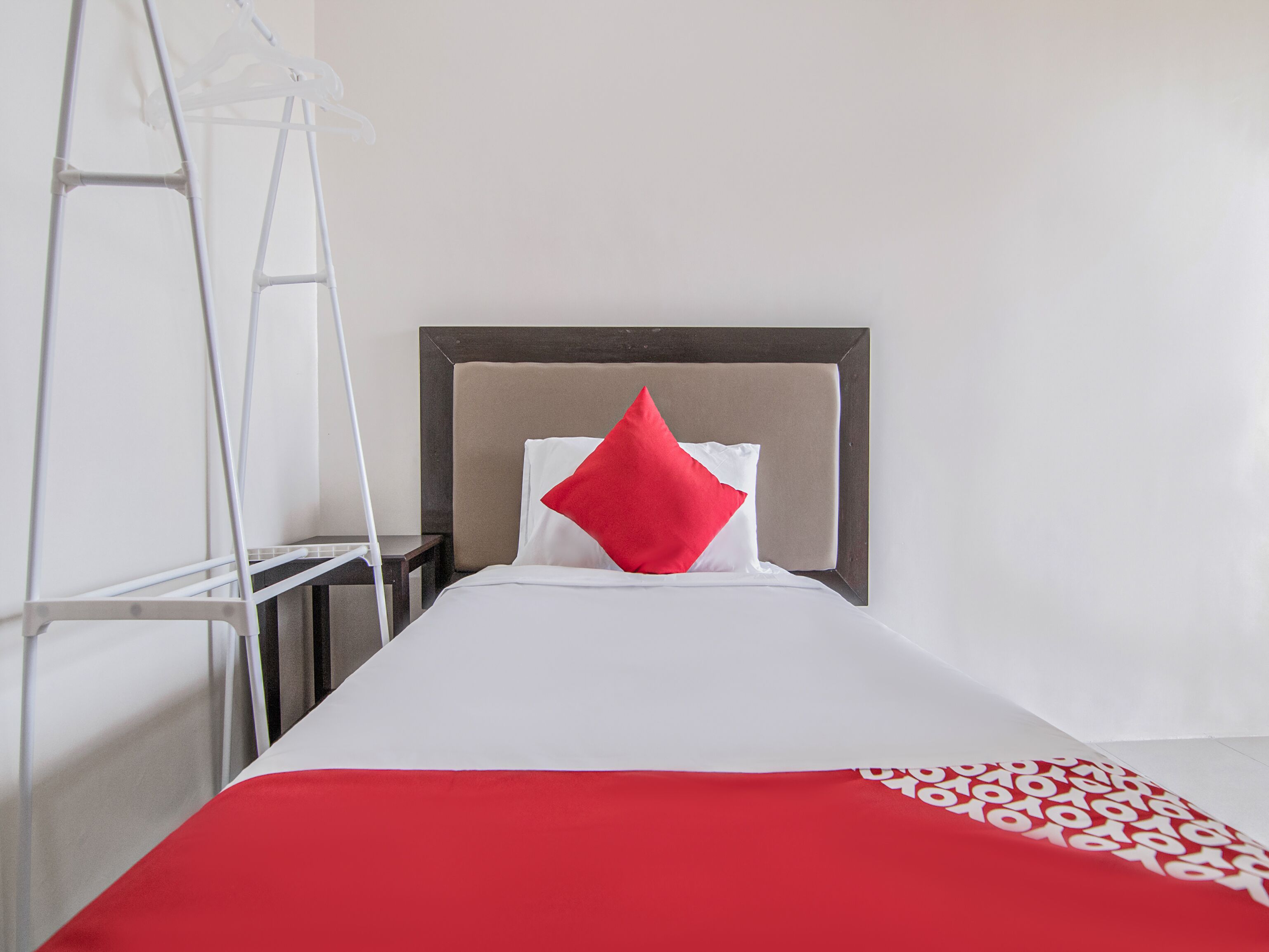Una cama bien hecha con un cojín rojo y una manta blanca, cabecera de madera y una escalera de color blanco apoyada en la pared.