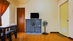 A room with a wooden floor, a television on a shelf, a standing fan, and a door. - OYO 431 Apollo 11 Apartelle (Pasay)