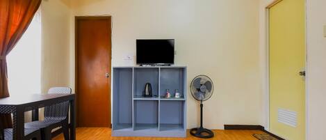 A room with a wooden floor, a television on a shelf, a standing fan, and a door.