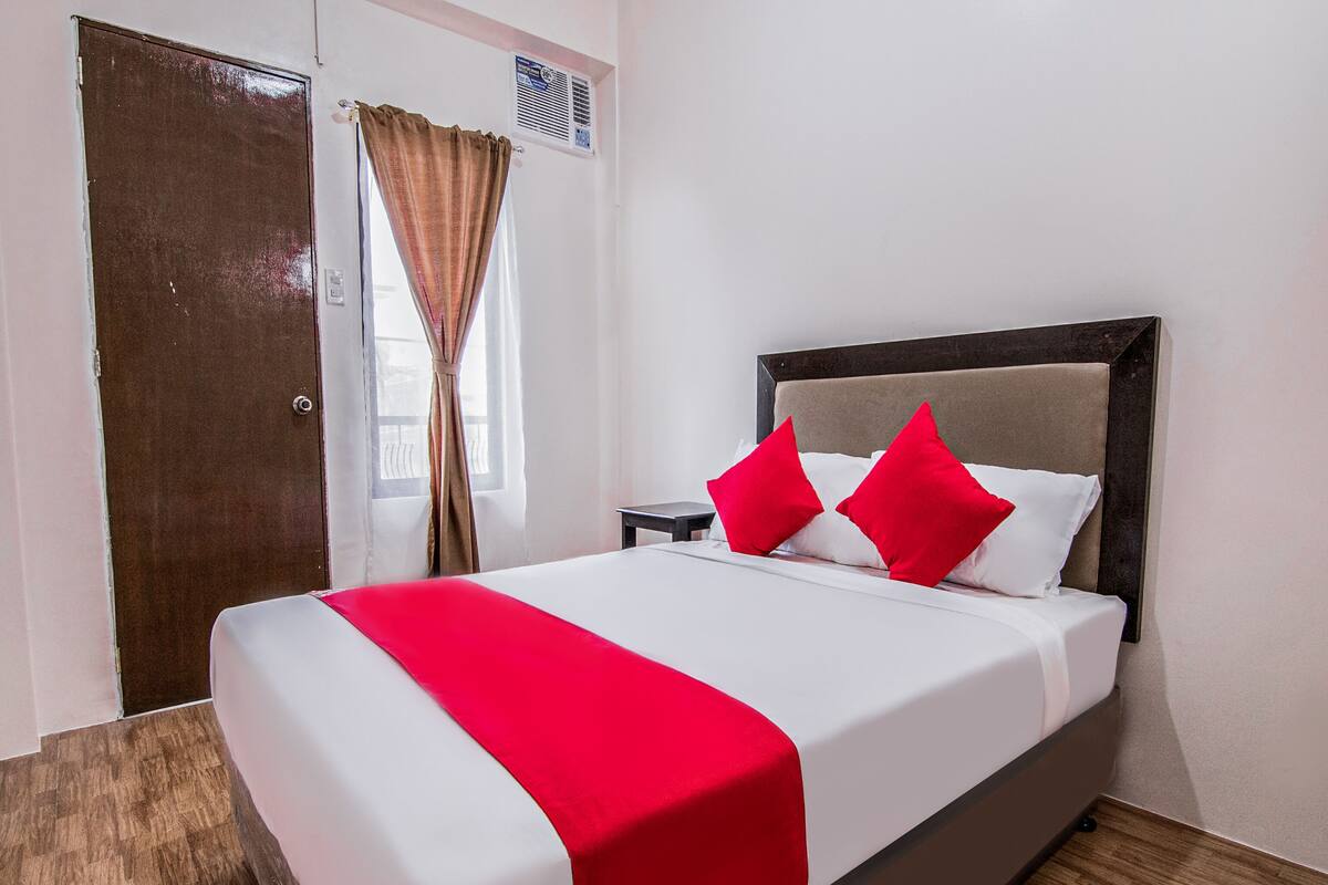 A hotel room with a bed, a nightstand, a window with curtains, and a door.