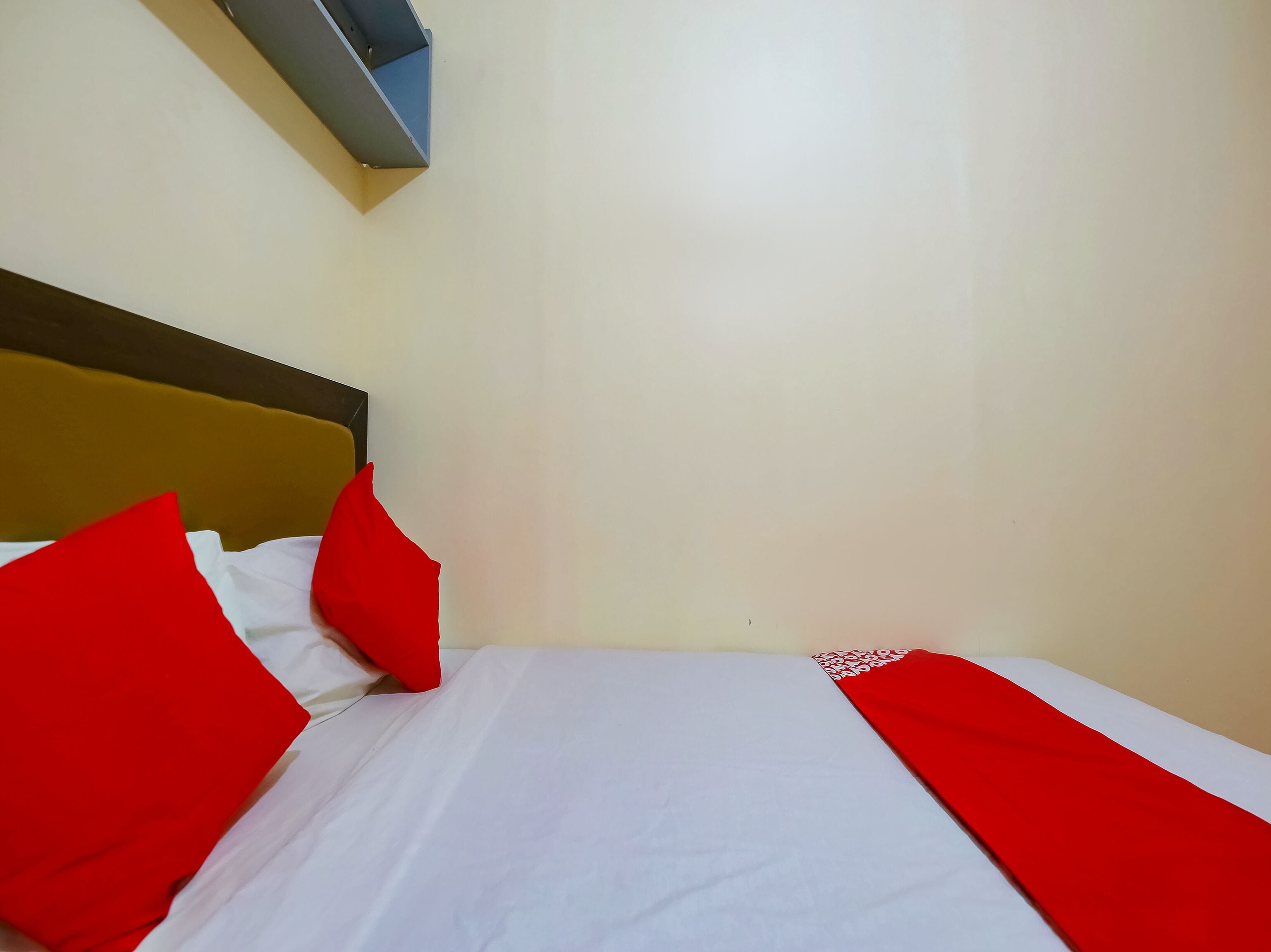 A bed with white sheets and red pillows, a wooden headboard, and a small blue shelf above it.