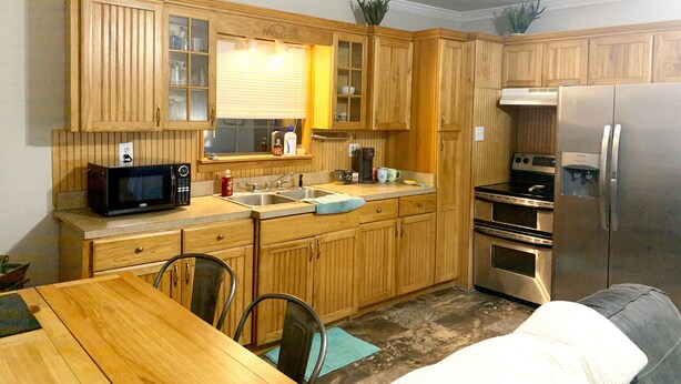 A compact kitchen with wooden cabinets, a microwave, a refrigerator, and a dining area with a table and chairs. - Pecan Orchard Bungalow (Purvis)