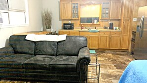A living room with a black leather sofa, a dining table, and a kitchen area with wooden cabinets and a refrigerator. - Pecan Orchard Bungalow (Purvis)