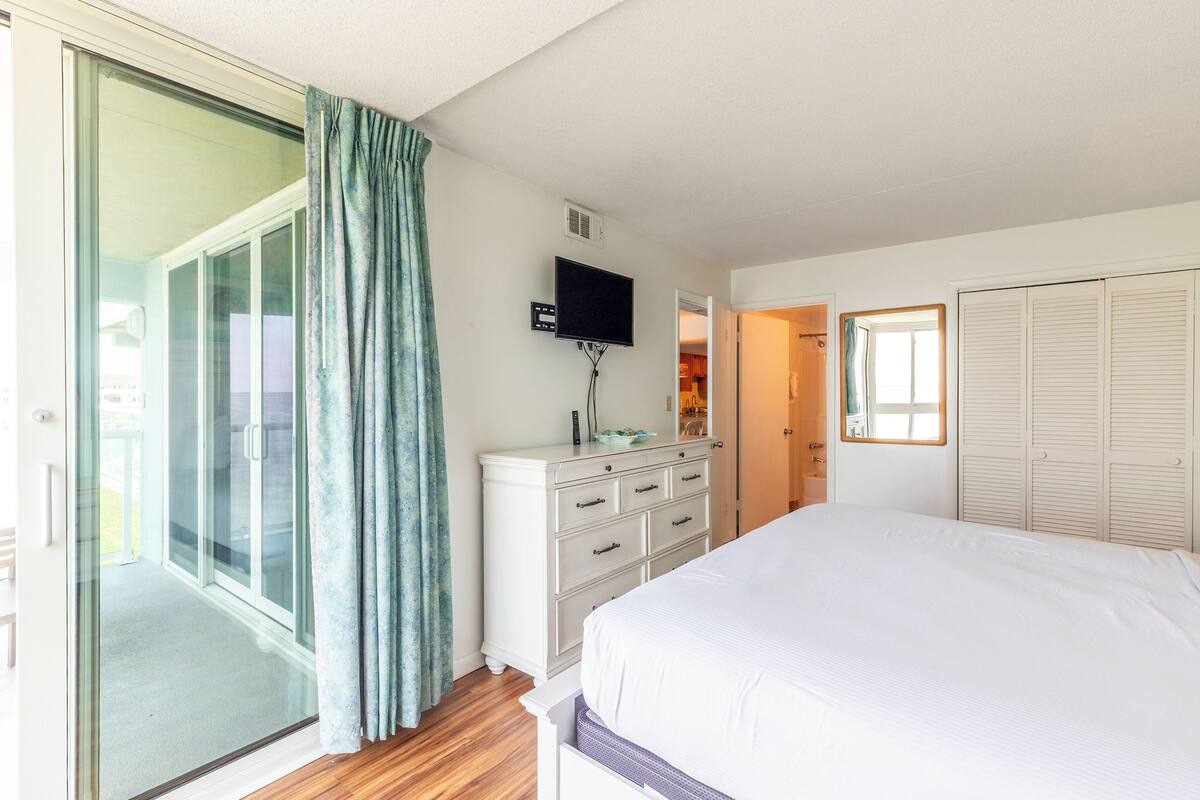 Ein Schlafzimmer mit einem Bett, einem Schminktisch, einem Fernseher und einer Schiebeglastür, die auf einen Balkon führt.