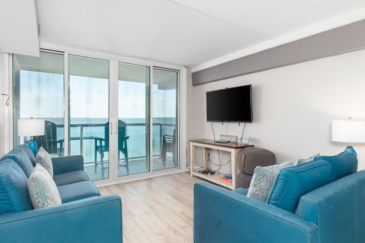 Ein modernes Wohnzimmer mit zwei blauen Sofas, einem Flachbildfernseher und einer Glastür, die auf einen Balkon mit Meerblick führt.