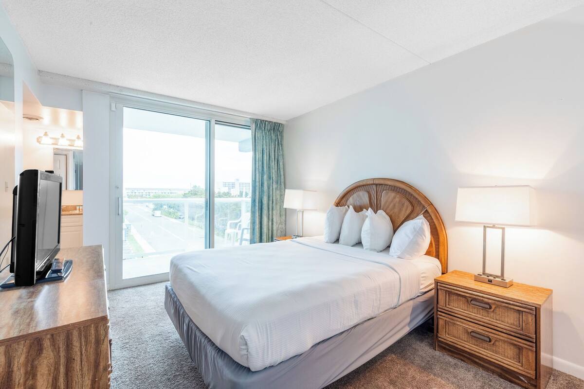 Ein Hotelzimmer mit einem großen Bett, einem Fernseher auf einer hölzernen Kommode, einem Nachttisch mit einer Lampe und Blick auf die Stadt durch ein großes Fenster.