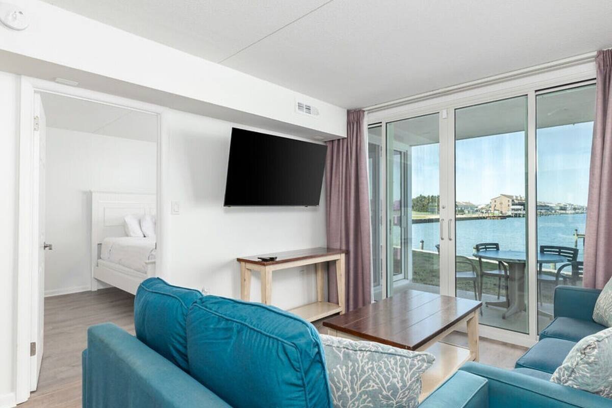 Ein modernes Wohnzimmer mit Flachbildfernseher, Sofa, Couchtisch und einer Schiebetür zu einem Essbereich im Freien mit Blick auf das Wasser.