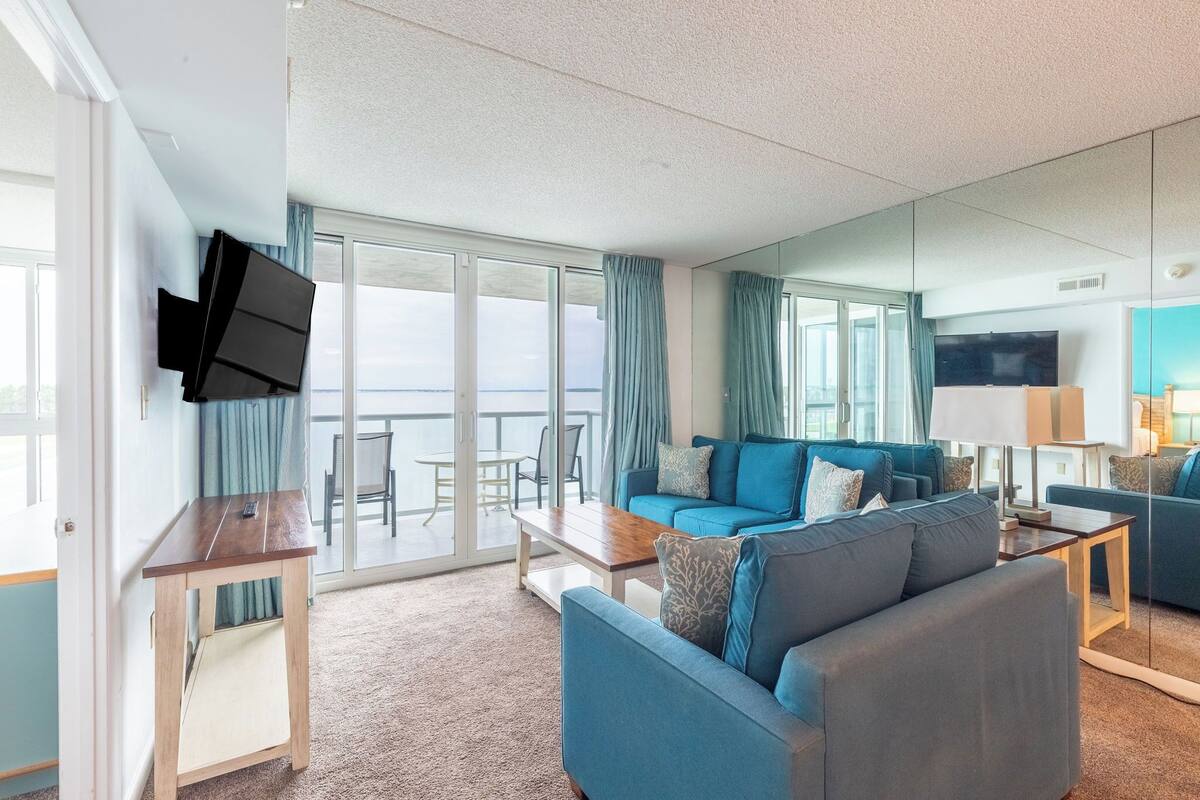Ein modernes Wohnzimmer mit Sofa, Couchtisch und Fernseher. Große Fenster bieten Blick auf das Meer.