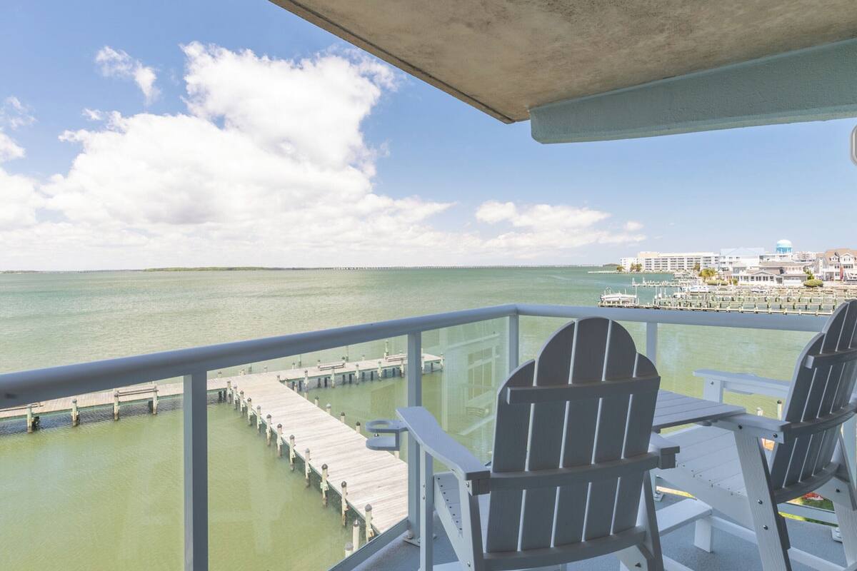 Ein Balkon mit zwei Adirondack-Stühlen, der auf einen Yachthafen und eine ferne Stadtansicht blickt.