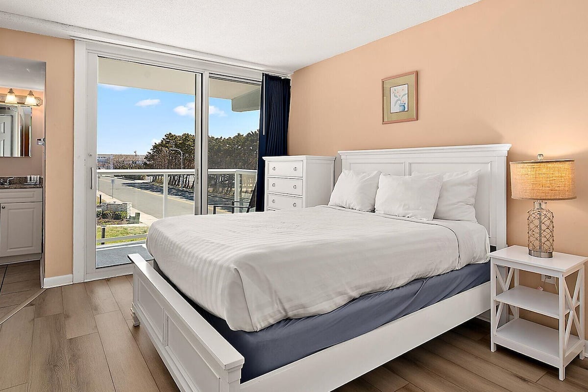 Ein Schlafzimmer mit einem großen Bett, einem Nachttisch mit Lampe, einer Kommode und einer Glasschiebetür, die auf einen Balkon führt.
