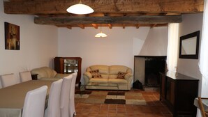 A cozy living room with a fireplace, a sofa, a dining table with chairs, and a wooden ceiling.
