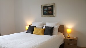 A neatly made bed with white and yellow pillows, a wooden nightstand with a lamp, and a framed picture on the wall.