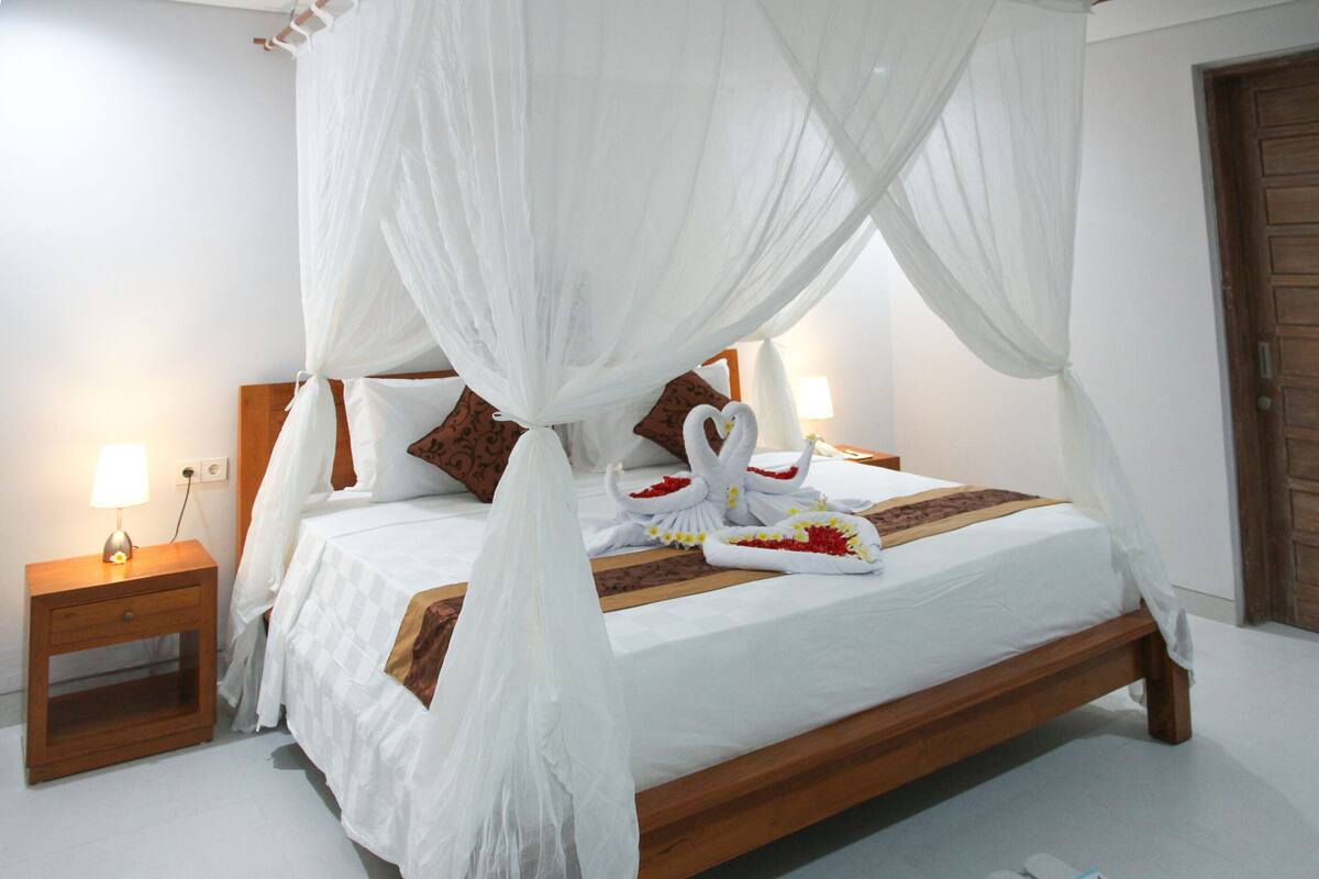 A four-poster bed with white bedding and a canopy, a wooden nightstand with a lamp, and a wooden door in the background.