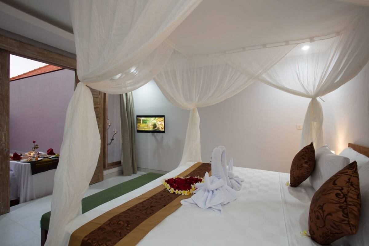 A room with a bed, white canopy, brown pillows, a TV, and a small table with a flower arrangement.