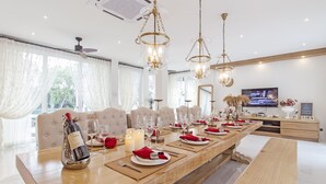 Uma sala de jantar com uma longa mesa de madeira posta para uma refeição, lustres e uma televisão ao fundo.