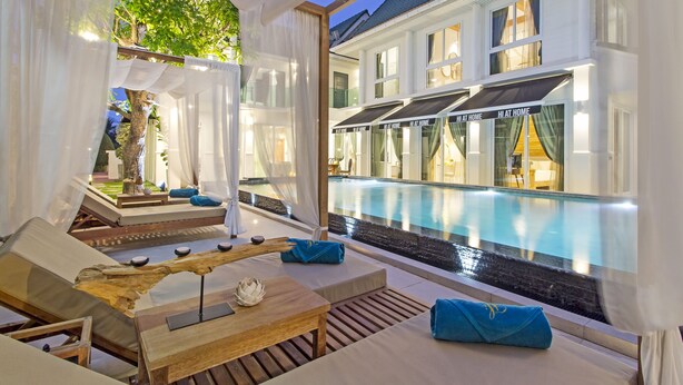 Uma área de descanso à beira da piscina, com espreguiçadeiras, uma mesa de madeira e vista para a piscina e o edifício do hotel.