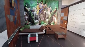 A modern lobby with a mural of a mythological scene, a brown sofa, a glass coffee table, and a wooden side table. - Hotel Rashika Palace (Jaipur)