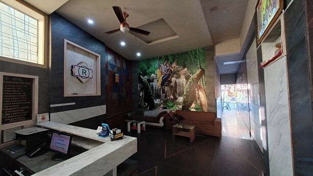 A hotel reception area with a reception desk, a ceiling fan, a wall-mounted sign with an 'R', and a mural of a waterfall. - Hotel Rashika Palace (Jaipur)