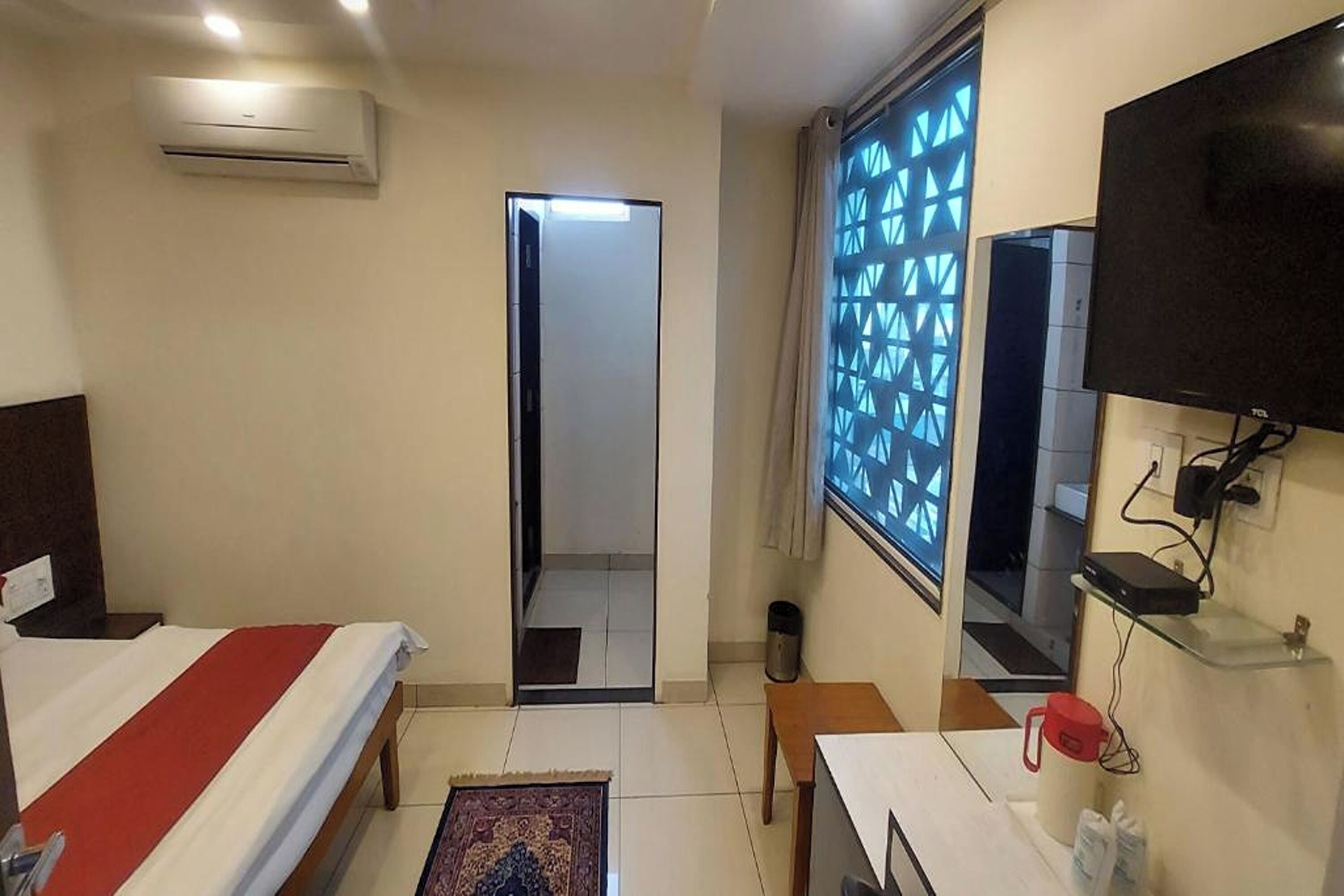 A hotel room with a bed, a TV, an air conditioner, a window with a geometric pattern, and a door leading to another room.