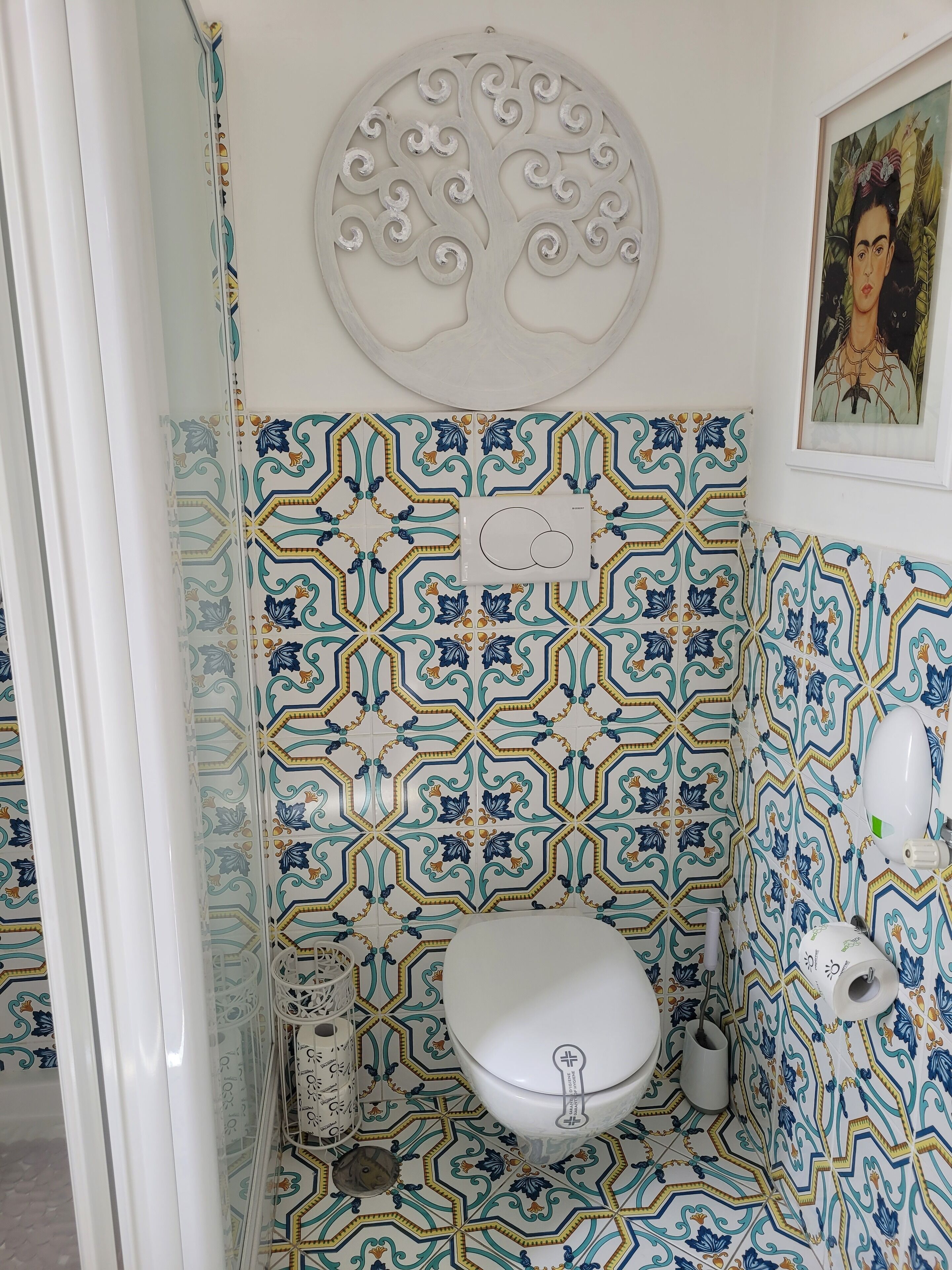 Een badkamer met een wit toilet, patroontegels, een decoratief wandobject en een lijst met een afbeelding.