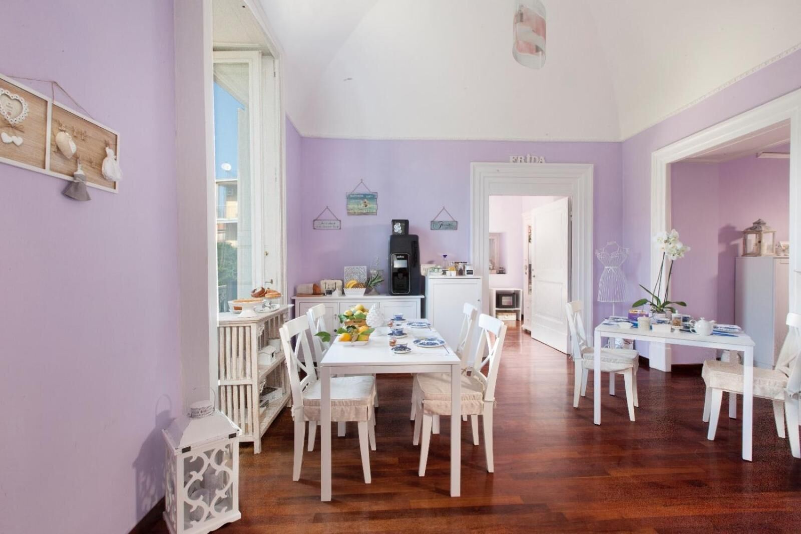 A dining area with white furniture, a kitchen in the background, and a purple wall.