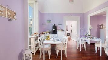 A dining area with white furniture, a kitchen in the background, and a purple wall.