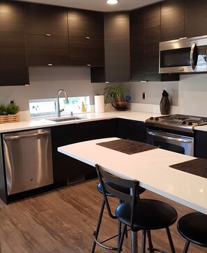 A modern kitchen with dark cabinetry, stainless steel appliances, a white countertop, and a dining area with two chairs.